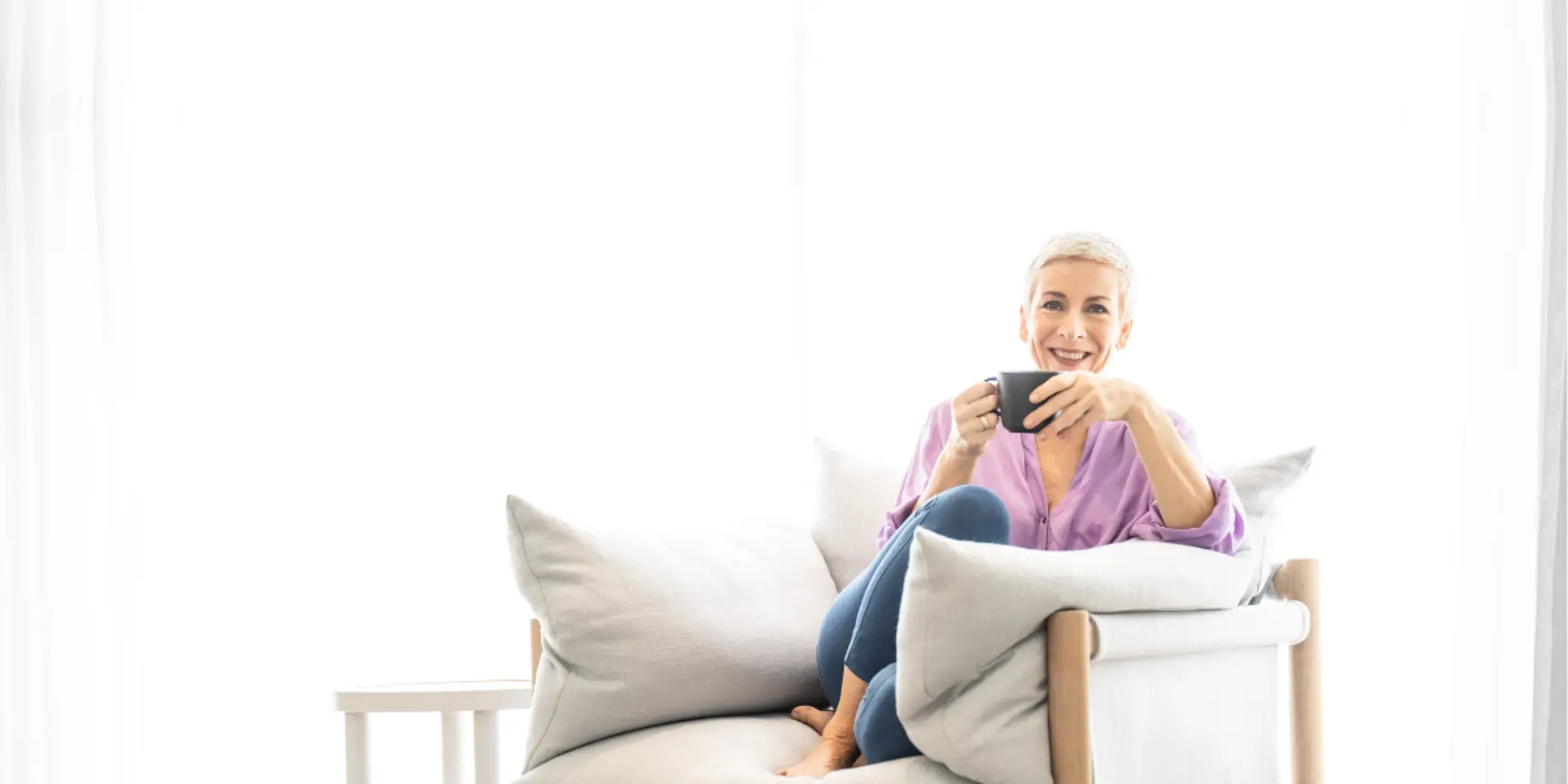 A woman curled-up on a chair with lots of cushions, holding a mug near her face.