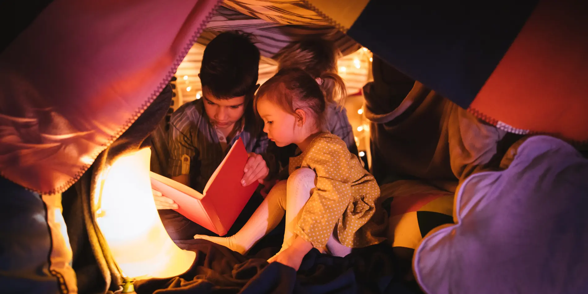 Three children reading a book while inside a blanket fort.