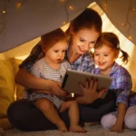 A woman holding a tablet and reading to two small children. They are all sitting inside a cubby made from a sheet.