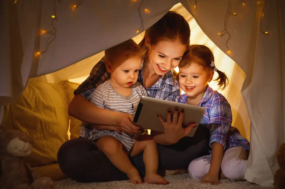 A woman holding a tablet and reading to two small children.