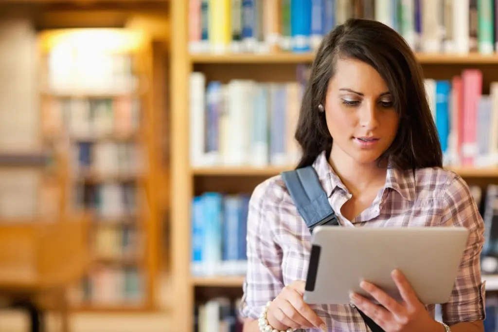 A woman standing in a library, looking at a tablet.