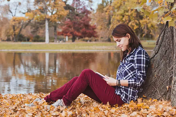 Woman reading a book under a tree, by a lake.