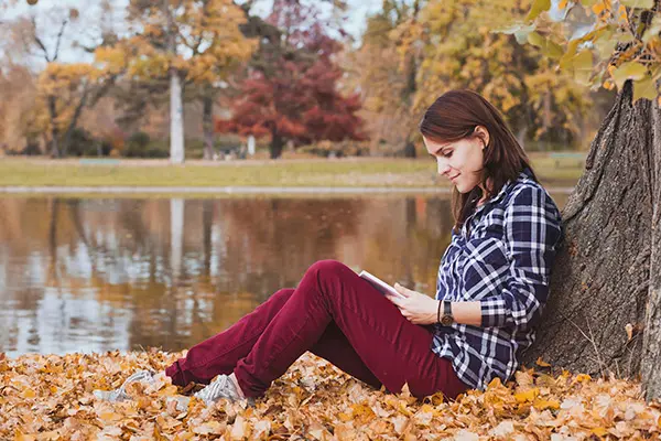 A woman reading under a tree, beside a lake.