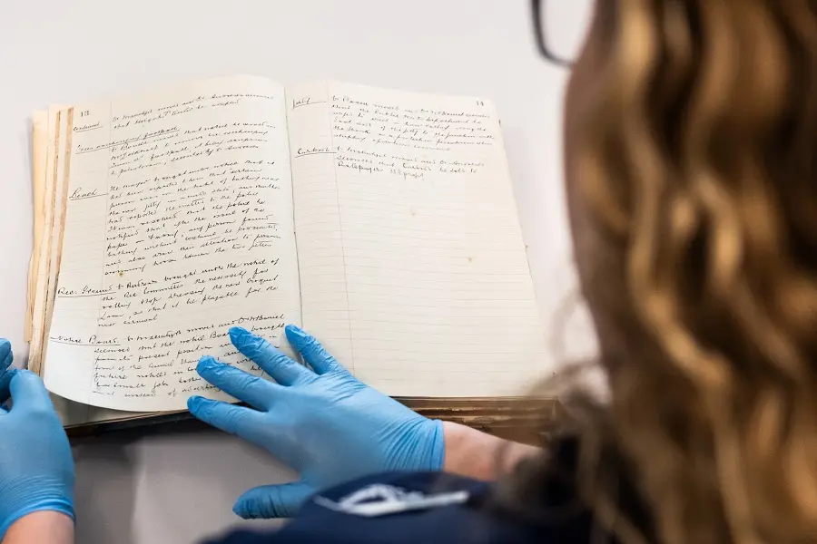 An old book viewed over the shoulder of a woman wearing gloves.