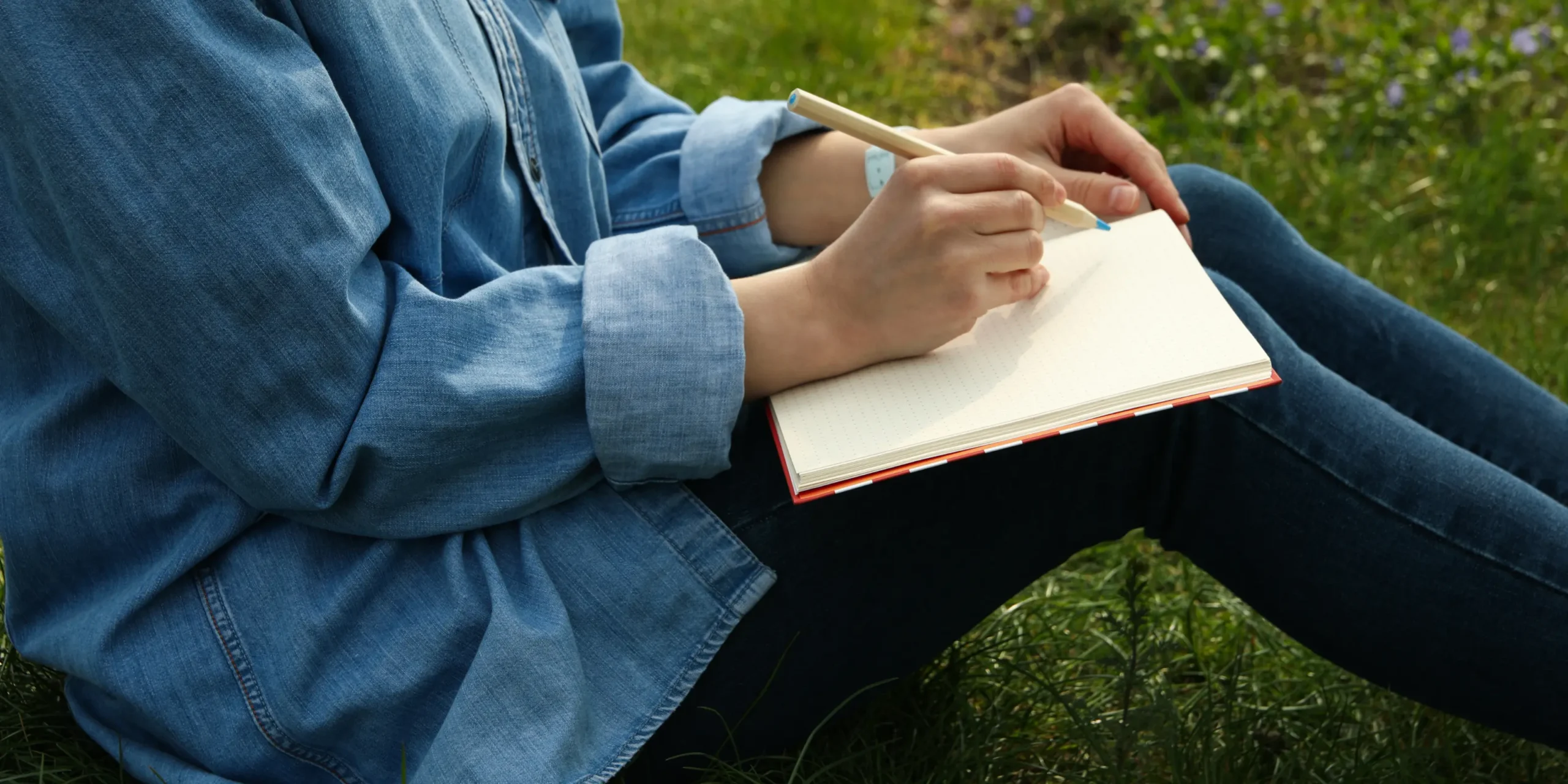 A close up of a person writing in a journal, sitting on grass.