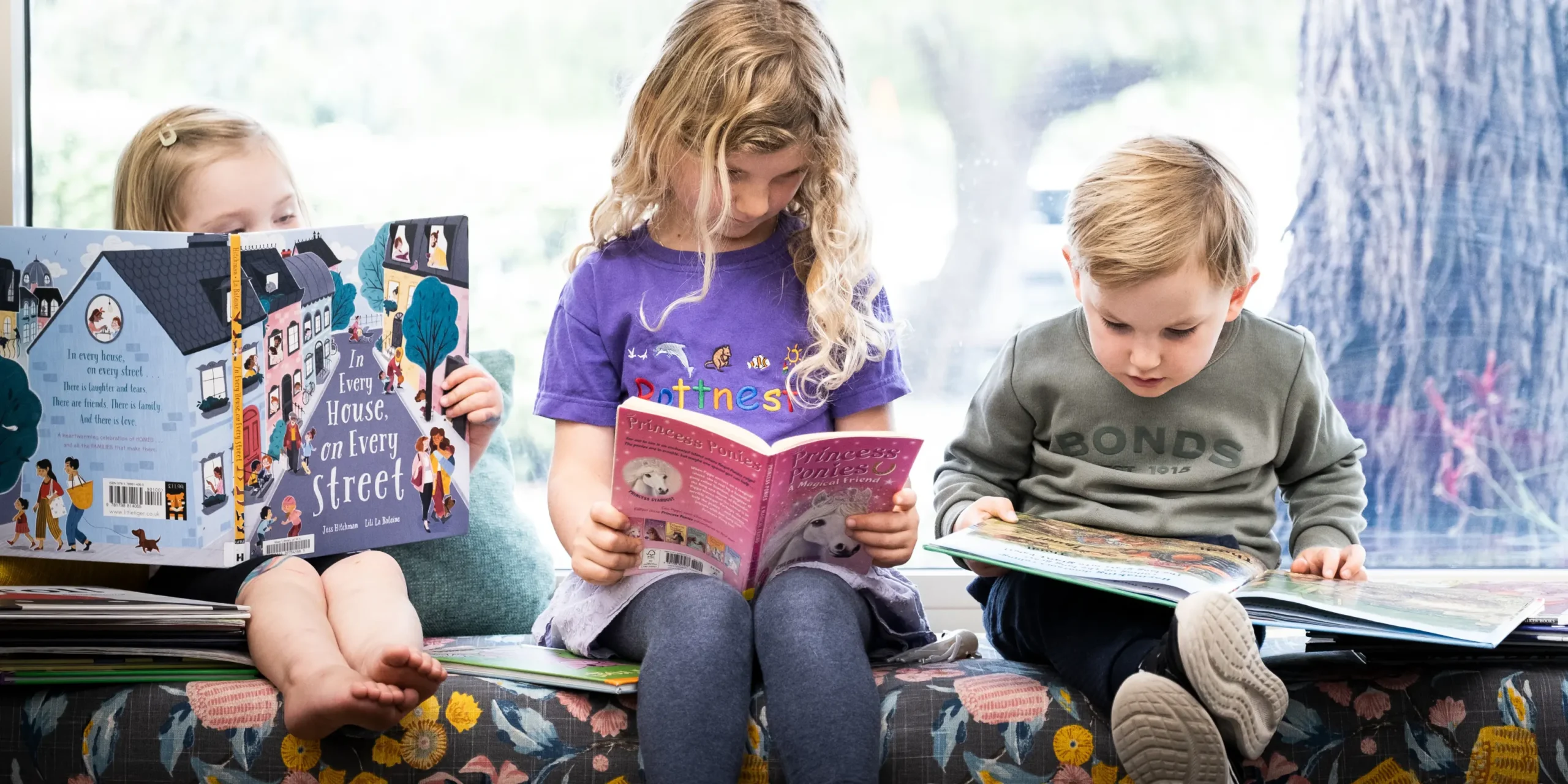 Three children sit on an indoor bench at the library, immersed in their books.