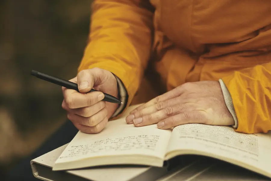 writers-group-900×600 A person, mostly out of shot, wearing a yellow jacket. They are holding an open notebook and a pen.