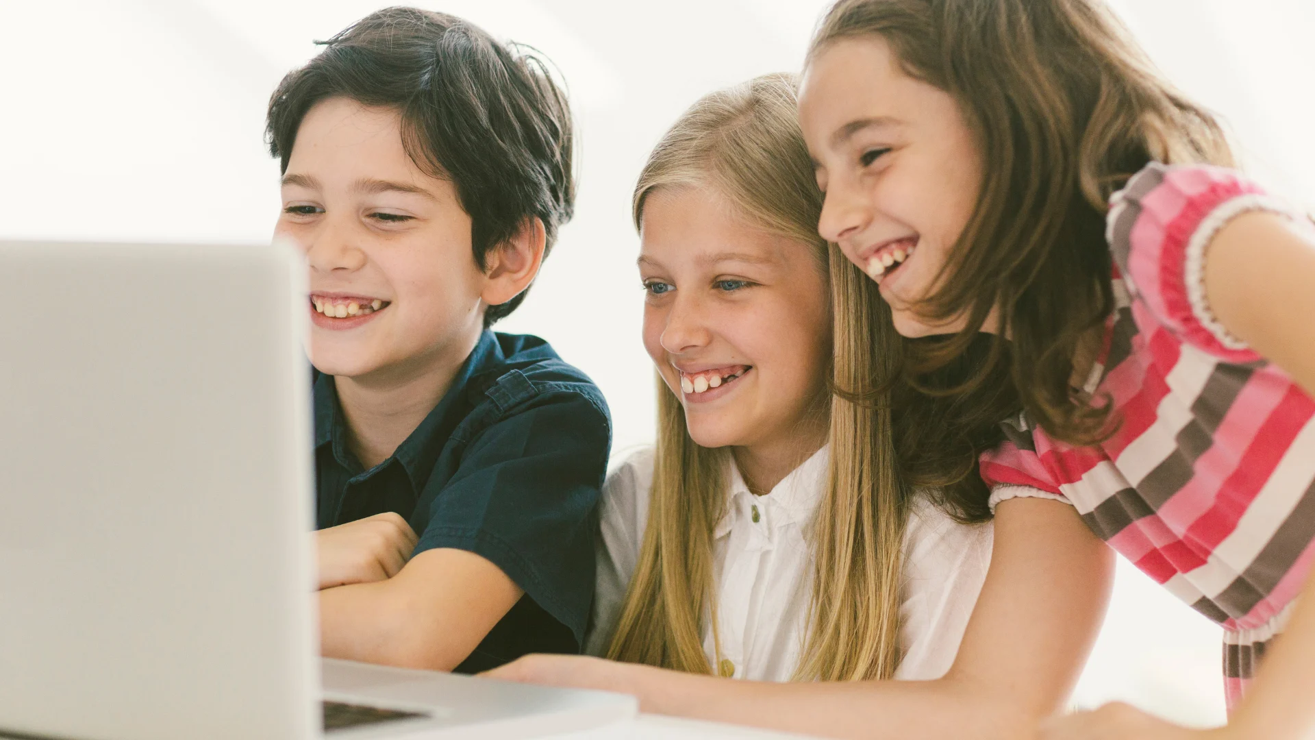 Three pre-teen kids surround a computer, smiling.