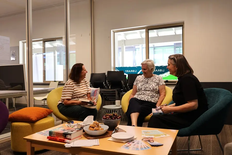 community-space Three women sitting in chairs in the Community Space. There are books on the table beside them.