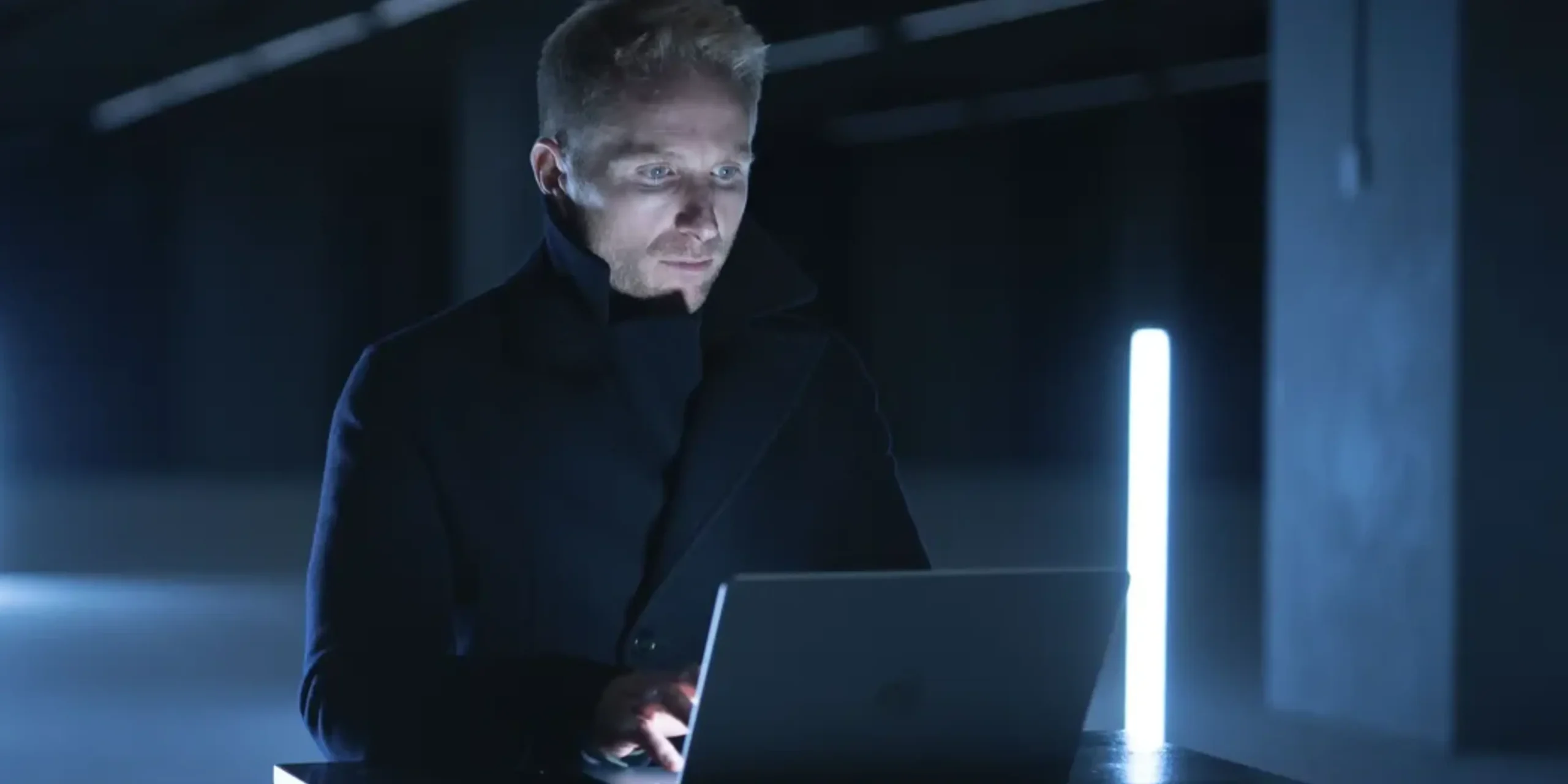 An image of a man sitting at a desk with a laptop, in a dark room.