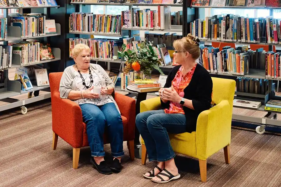 Two women sitting in the library, holding mugs and chatting with each other.