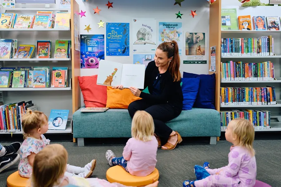 storytime-2026 A woman sitting in the middle, reading a book to a group of children sitting in front of her.