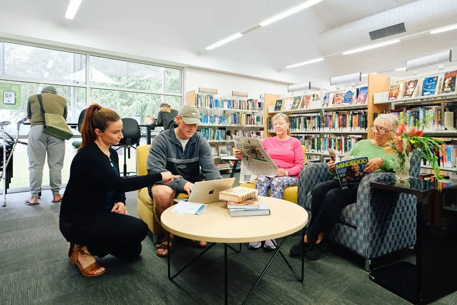 Three people sitting around a table. Two are reading, while a third has a laptop and is receiving assistance from a librarian.